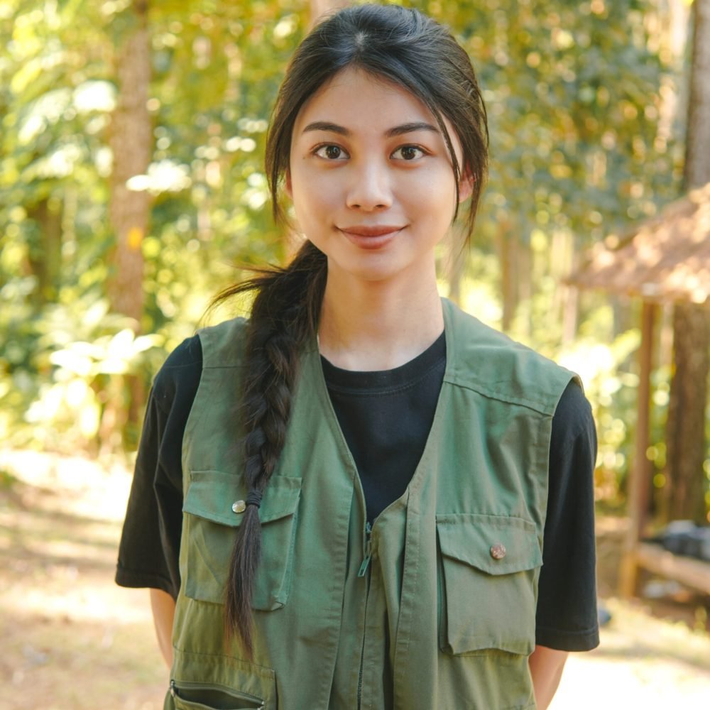 Confident young Asian woman wearing a green vest smiling to the camera with forest nature background.